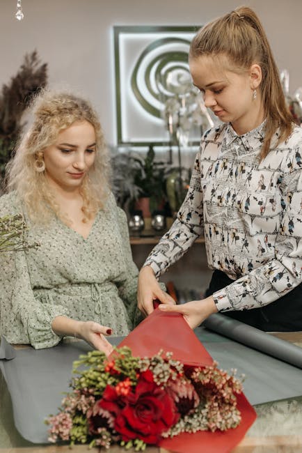 A florist with long red hair, dressed in a patterned blouse, arranging a floral bouquet wrapped in red paper on a work table inside a shop. The bouquet features large red roses, smaller pink flowers, 