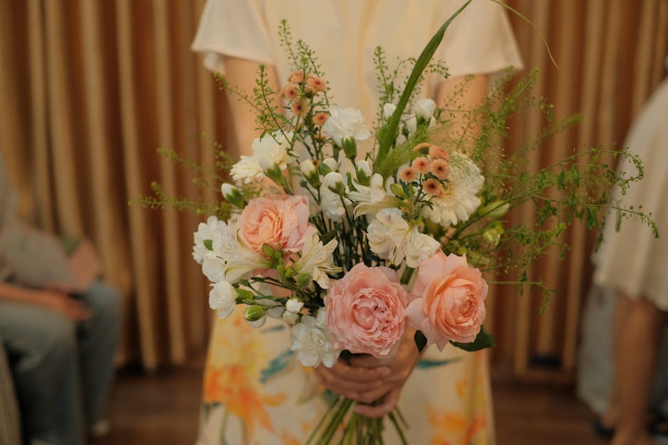 A person holding a floral bouquet featuring soft pink roses, white lilies, and small peach-colored spray chrysanthemums, complemented by delicate green foliage and ferns. The bouquet is arranged with 