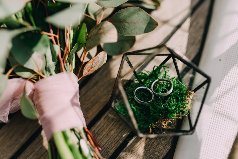 A close-up of a floral arrangement featuring a bouquet of eucalyptus leaves with rounded, greenish-gray foliage and reddish stems, wrapped in soft pink tissue paper. Beside the bouquet, there is a sma