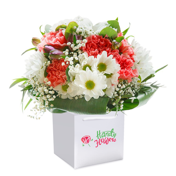 Arranged with finesse in a crisp square white gift bag, this exuberant bouquet captures the spirit of Mortlake's riverside charm, ready to bring a burst of colour to any address from Ship Lane to Avondale Road. The focal point is a lush medley of coral and peach carnations, each blossom presenting ruffled, velour-like petals and an irresistible sense of depth. Sprightly white daisies with vivid lime-yellow eyes rise between the carnations, their brightness echoing the lively lawns and flowerbeds of Mortlake Green. Airy clouds of baby's breath delicately float throughout, softening transitions and adding a gentle, nostalgic tone. Not to be missed among the arrangement's left edge are several closed buds with a subtle purple flush, adding dimension and hinting at blooms just about to open. Framing and supporting the vibrant cluster, broad, glossy green leaves curl in wide arcs, imparting structure and a sense of fullness. The carefully rounded dome shape reflects an expert's touch, balancing cheerfulness and refinement. The modern gift bag-its white ribbon handles impeccably tied-suggests ease of carrying and a spirit of celebration. The bouquet's poise against a spotless white backdrop accentuates its colours and freshness, making it an ideal offering for birthdays, anniversaries, or spontaneous gifts sent throughout Mortlake, Barnes, or East Sheen. Every flower and leaf has been thoughtfully chosen and placed, reflecting both local flair and timeless elegance.
